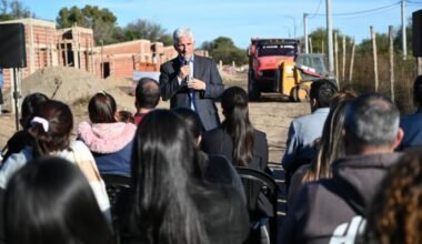 El gobernador Claudio Poggi habla ante vecinos durante una recorrida por el predio de las viviendas en construcción, con casas de ladrillo y maquinaria al fondo.