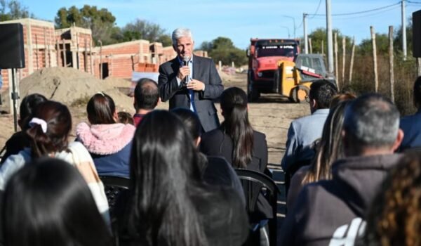 El gobernador Claudio Poggi habla ante vecinos durante una recorrida por el predio de las viviendas en construcción, con casas de ladrillo y maquinaria al fondo.