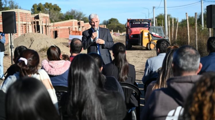El gobernador Claudio Poggi habla ante vecinos durante una recorrida por el predio de las viviendas en construcción, con casas de ladrillo y maquinaria al fondo.